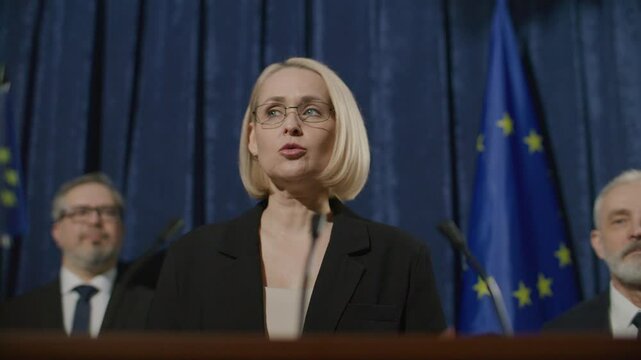 Middle-aged female diplomat wearing glasses and formal suit, speaking at podium during political event with European Union flags and blue curtain background, flanked by two male colleagues