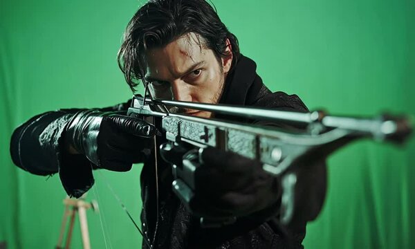 A determined man aims a crossbow directly at the viewer with a stern expression on his face.