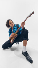 Fototapeta premium Young energetic man in tropical shirt, sitting and passionately playing electric guitar against white background. Concept of music, performance, festival, talent, passion, artistic movement.