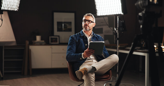 Live stream host, talk show and man with tablet in home studio for documentary interview or online podcast. Filming set, lights and mature journalist with digital script for recording and news update