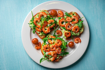 Toast with shrimp, arugula and avocado on a white plate with cherry tomatoes on a blue table. Space for text. Healthy and tasty breakfast concept. Top view. Flat lay.