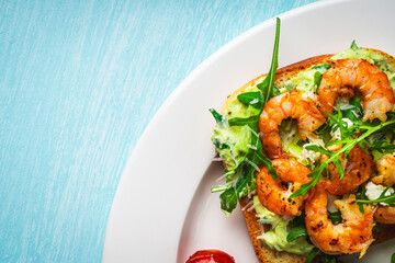 Toast with shrimp, arugula and avocado on a white plate with cherry tomatoes on a blue table. Space for text. Healthy and tasty breakfast concept. Top view. Flat lay.