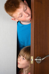The children, a brother and a sister, peek curiously from behind the door of the room. vertical photo