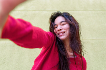 Fototapeta premium Young woman taking selfie, wearing red sweater against green wall backdrop, long brown hair framing face