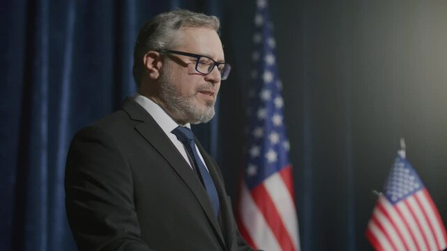 Middle-aged senator in formal suit delivering political speech from stage with American flags in background during press conference. Tilt-up shot