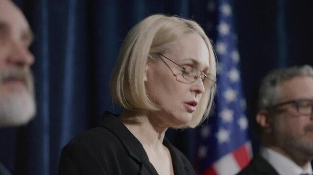 Female senator standing on stage with two male colleagues, delivering speech to audience during political press conference with US flag in background
