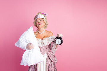 Elegant woman in historical costume holding pillow and vintage clock against a soft pink background