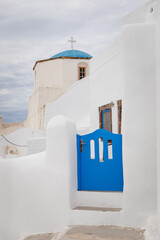 Vertical view of perfect holiday apartment with blue door and gate in Pyrgos Kallistis in the island of Santorini, Greece, a Greek village in mainland with an orthodox church, cross, and cloudy sky