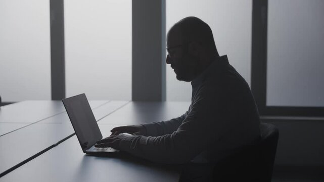 Man fertigt Bericht im B&uuml;ro an Schreibtisch auf Laptop an, sitzt im Schatten mit Gegenlicht der Fenster