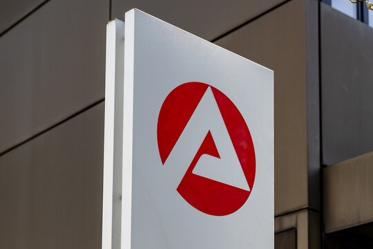Nahaufnahme eines Schildes mit dem Logo der Bundesagentur f&uuml;r Arbeit. Das rote Symbol auf wei&szlig;em Hintergrund steht vor dem Geb&auml;ude der Arbeitsagentur.