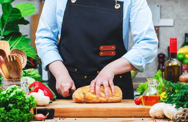 Chef in apron cuts fresh ​​ciabatta bread. Cozy kitchen with wooden table, kitchenware and ingredients for cooking. Homemade baking, cooking, food blogging