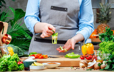 Cook holds core of green paprika. Cozy kitchen with wooden table, kitchenware, vegetables, herbs and ingredients for cooking. Healthy vegan eating, culinary, recipes, food blogging