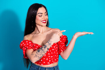 Smiling young woman in red floral top pointing hand on blue background showcasing casual fashion...
