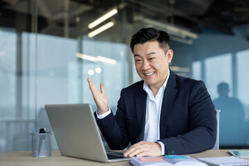 Happy young Asian man in a business suit sitting at a desk in an office, looking at a laptop screen and rejoicing in success and news