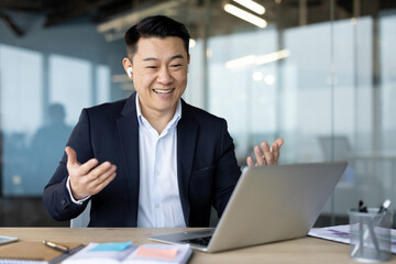 Asian smiling young man in a suit and headphones sitting in the office in front of a laptop and talking on video call gesturing with his hands
