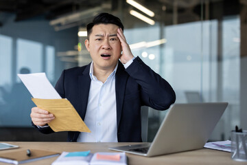 Upset young Asian businessman sitting in the office at his desk, holding an open envelope with a...