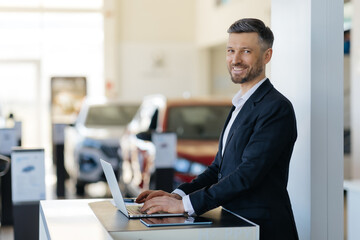 Professional car salesman in black suit standing confidently at modern car dealership workstation,...