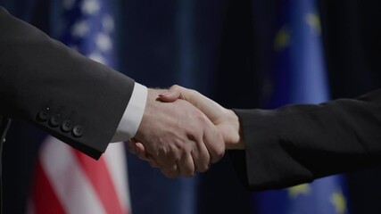 Close-up of handshake between male and female politicians in formal suits during international press conference, with American and European Union flags in background - Powered by Adobe