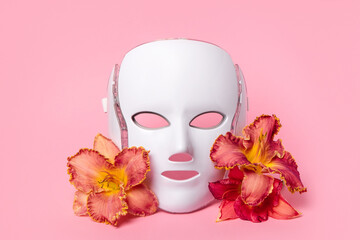 Cosmetic LED facial mask regenerative treatment on pink background with flowers lilies. Health and...