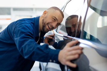 Cheerful man embracing his new car with excitement inside modern dealership showroom. The polished...