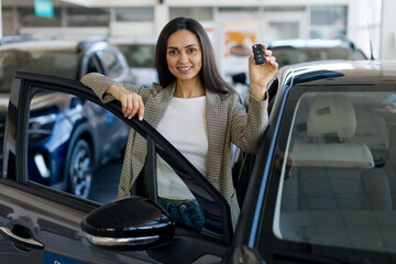Happy woman standing next to his newly purchased car at modern dealership showroom, holding up the...