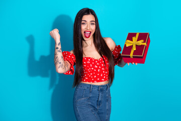 Excited young woman holding a gift box, dressed in trendy outfit, standing against a bright blue...
