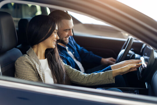 Young Customers Couple Sitting In Car Pushing Buttons Testing Auto In Dealership Showroom, Millennial Family Buying Their New Automobile, Happy Man And Woman Examining Vehicle Features