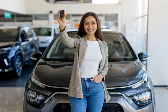 Happy woman standing in front of car in huge auto showroom, showing key from automobile and smiling at camera, millennial female got new vehicle at dealership center , copy space