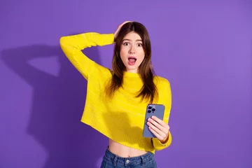  Surprised young woman with smartphone making expressive gesture on purple backdrop, representing communication and lifestyle fashion © deagreez
