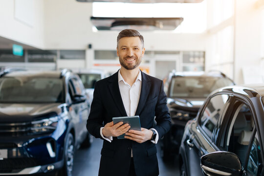 A professional car salesman in black suit and white shirt stands confidently in modern car dealership showroom filled with luxury vehicles, man smiling while holding digital tablet, looking at camera