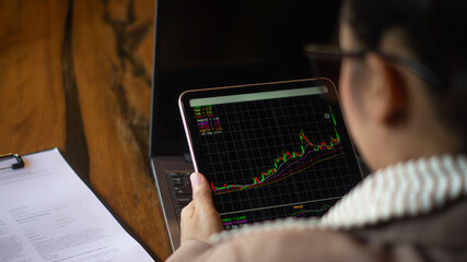 Young female investor looks at tablet in hand, on screen shows candlestick chart of stock price, businesswoman manages investment plan at desk with laptop and checks documents.