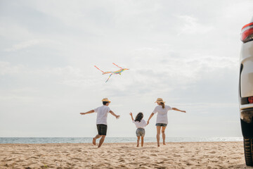 Asian family is flying a kite while running on the beach. Happy parents mother and father with their child playing with kite, The car is parked nearby, and the family is enjoying their time together