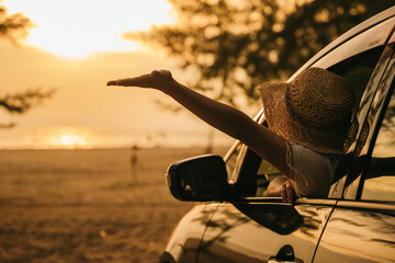 Woman wearing straw hat sits in front seat waving hand out car window at sunset feeling the wind. Happy weekend travel enjoy summer at beach. summer vacation travel driving road trip concept