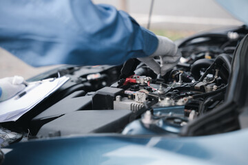 Focused Asian mechanic with a clipboard inspecting car engine in garage. Expertise in car service...