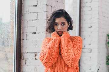 Young woman in an orange sweater standing near a window, exuding cozy and relaxed vibes in a...