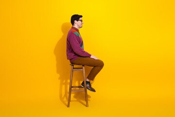 Young man sitting on stool against bright yellow background wearing fashionable casual attire and glasses, showcasing style and confidence.