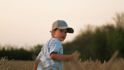 Cheerful child kid happily running through wheat field, turning around, looking at camera. Merry...