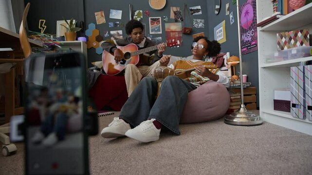Rack focus of creative Black teenage girl and boy recording vlog on smartphone on tripod while singing and playing ukulele and guitar in cozy room with maximalist interior at home