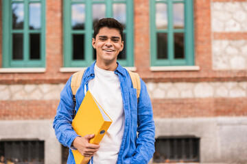Smiling university student holding a yellow binder in front of the university building