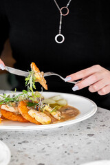 A plate with chicken, hash browns, garnished with fresh microgreens, served with pickles and sauce. A person is cutting the meal, showcasing a close-up dining moment.