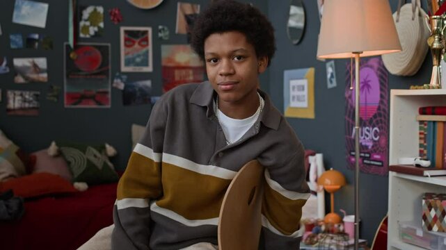 Tilt up slowmo portrait of African American teenage boy in casualwear looking at camera sitting on chair in his room with lots of posters and books