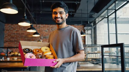 Young man in a casual outfit holding a pink box of assorted donuts in a cozy bakery, warm lighting and inviting atmosphere.
 - Powered by Adobe