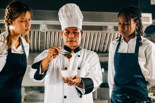Senior Asian chef mentors multiracial students in a restaurant kitchen workshop emphasizing teamwork learning and note-taking. Professional education in action. Food Edocation