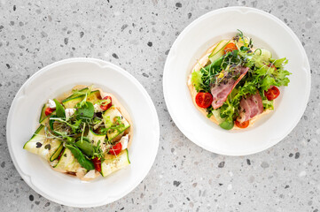 Two fresh salads on white plates atop a terrazzo countertop. One with zucchini ribbons, tomatoes,...