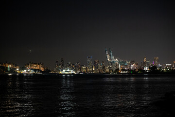City skyline illuminated at night with boats on the water reflecting vibrant lights in a bustling urban environment