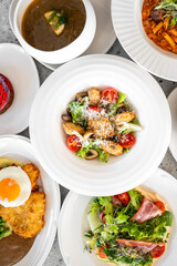 Top view of a variety of colorful dishes, including soup, pasta, salad, fried chicken, a fried egg, breaded meat, and dessert, beautifully presented on a table.