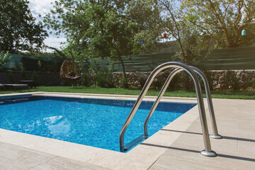 A small private swimming pool with a metal ladder is surrounded by greenery, a stone fence, and a...