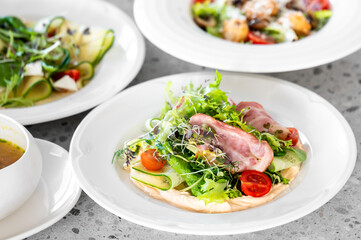 A fresh mixed salad featuring cherry tomatoes, cucumber ribbons, and ham slices on a white plate, with other dishes in the background. Perfect for healthy eating concepts.