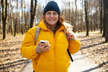 A happy woman in a yellow jacket and black beanie walking in the park, holding her phone. The bright natural setting, with trees and a peaceful path, enhances the cheerful mood of the moment.