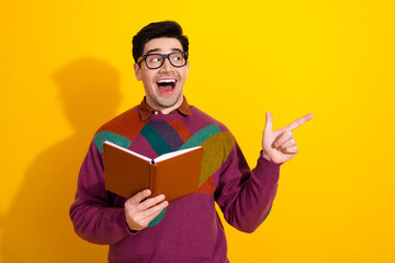 Excited young man wearing retro-style sweater holding a book and pointing with joy on a vibrant yellow background